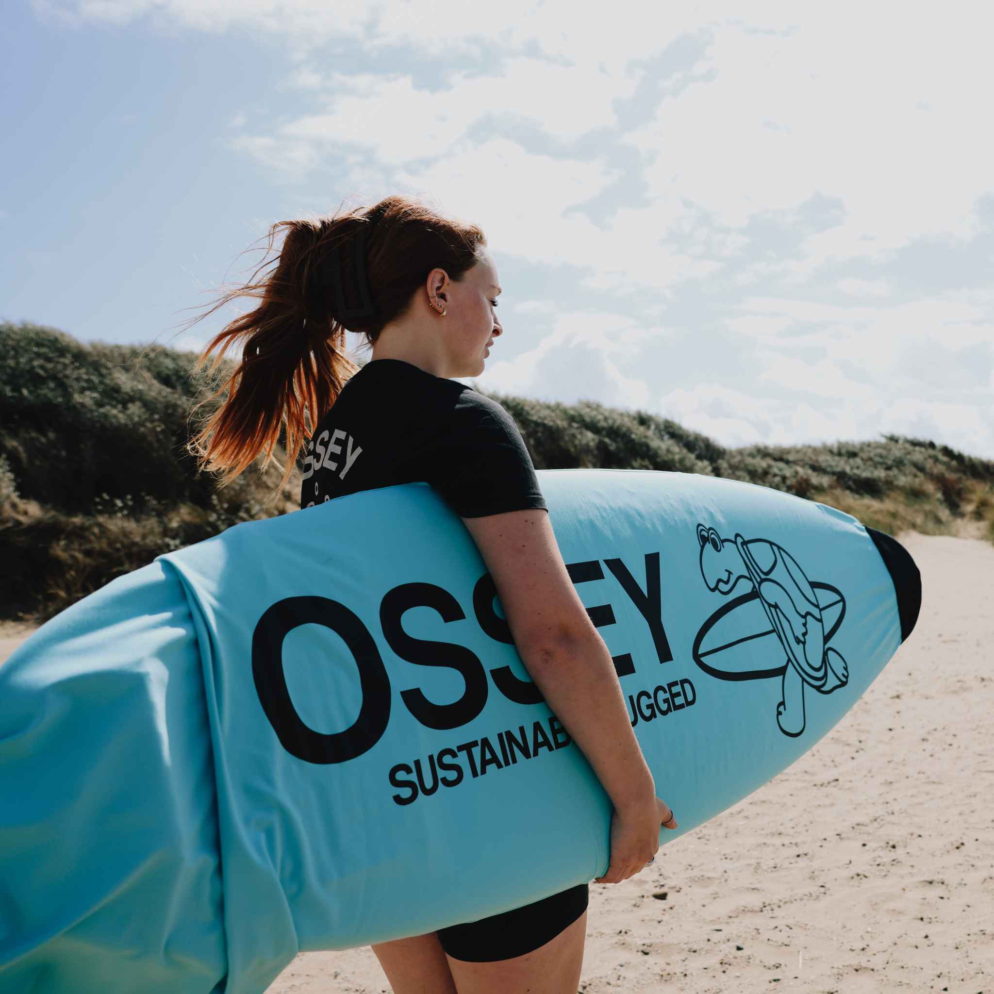 Person holding a blue surfboard sock with 'OSSEY' branding on a beach.