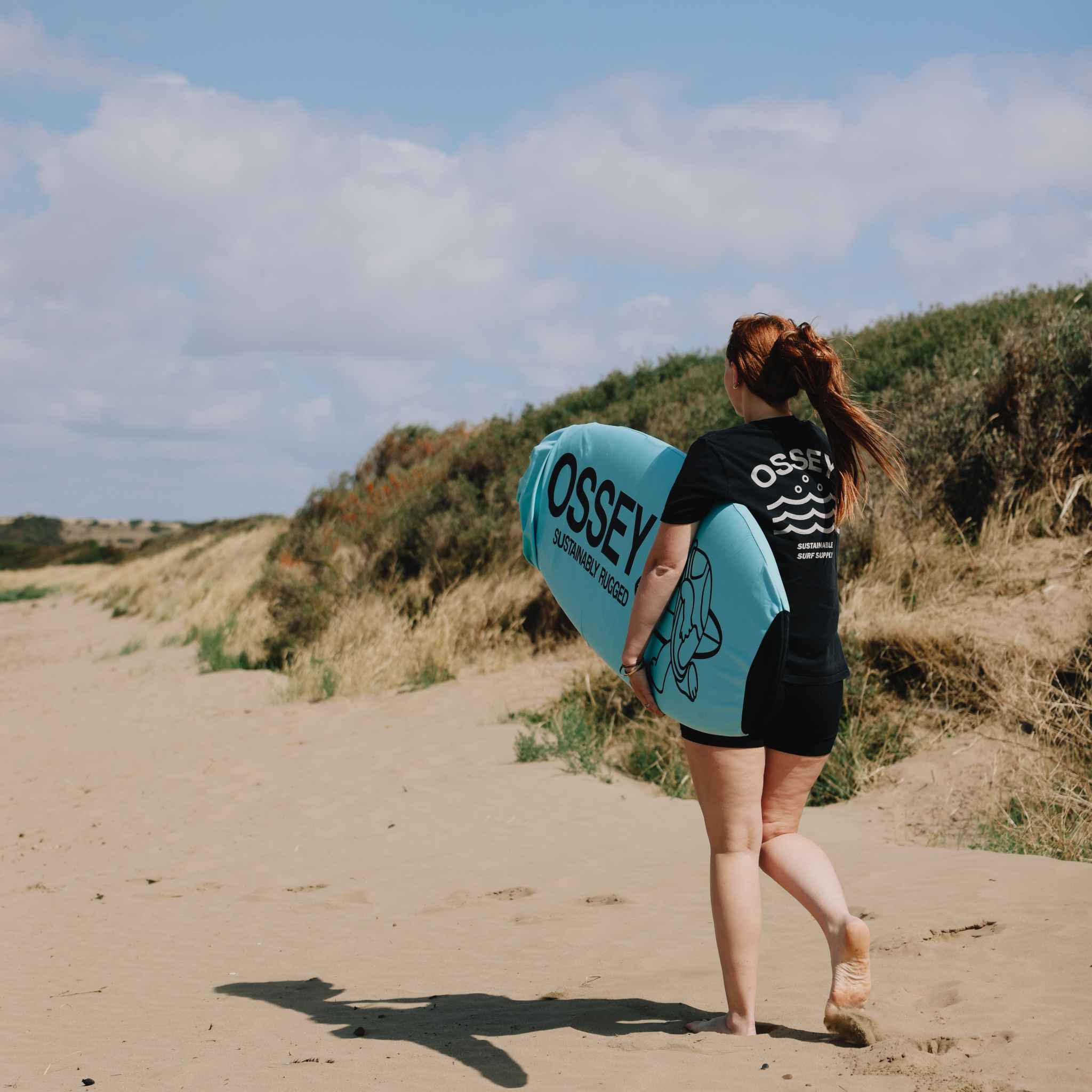 Person walking on a sandy path with a blue OSSEY surfboard sock