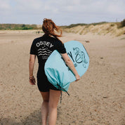 Person walking on a beach carrying a light blue surfboard sock with 'OSSEY' branding.