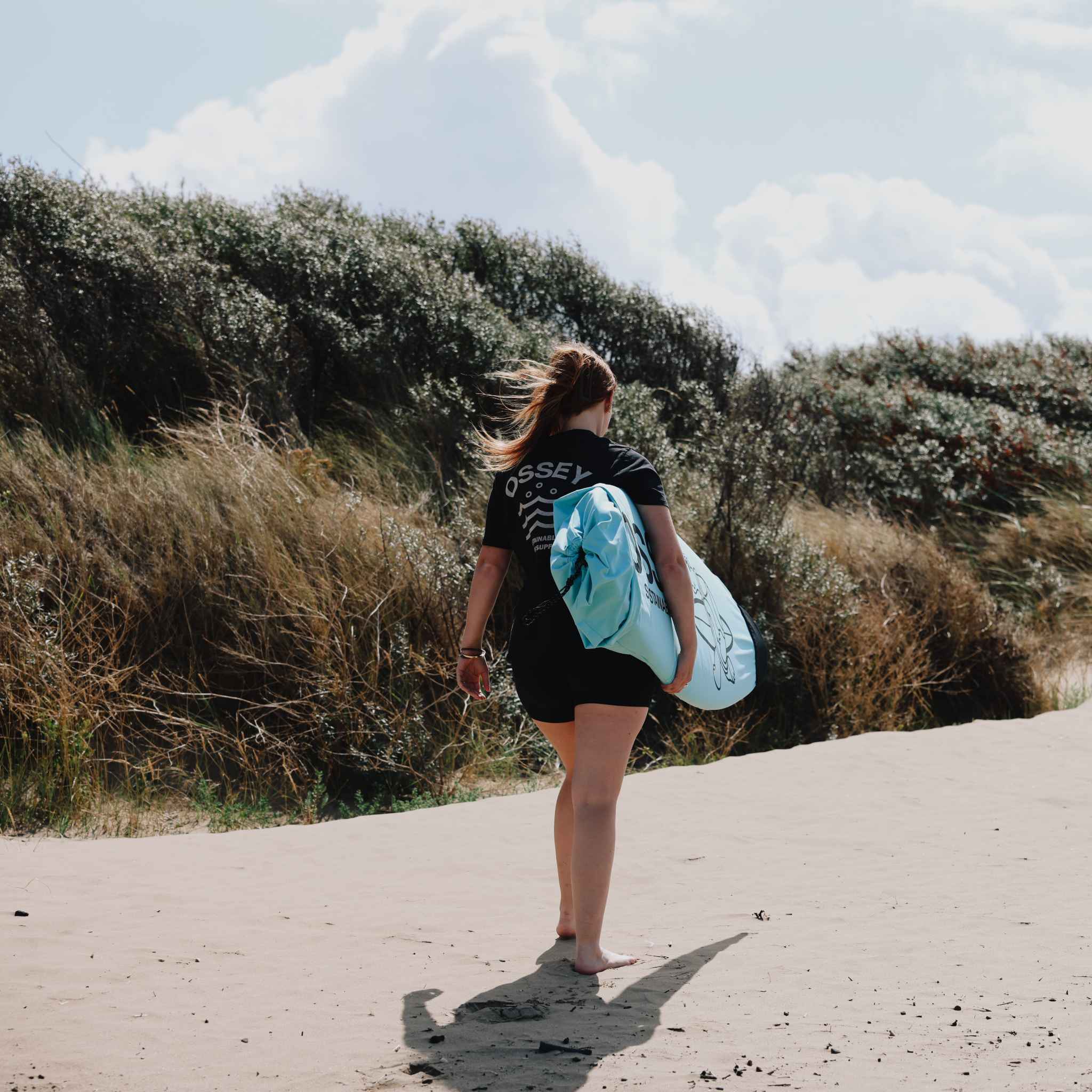 "Person walking on a sandy path with a surfboard sock under their arm, wearing a black t-shirt with 'OSSEY' branding."